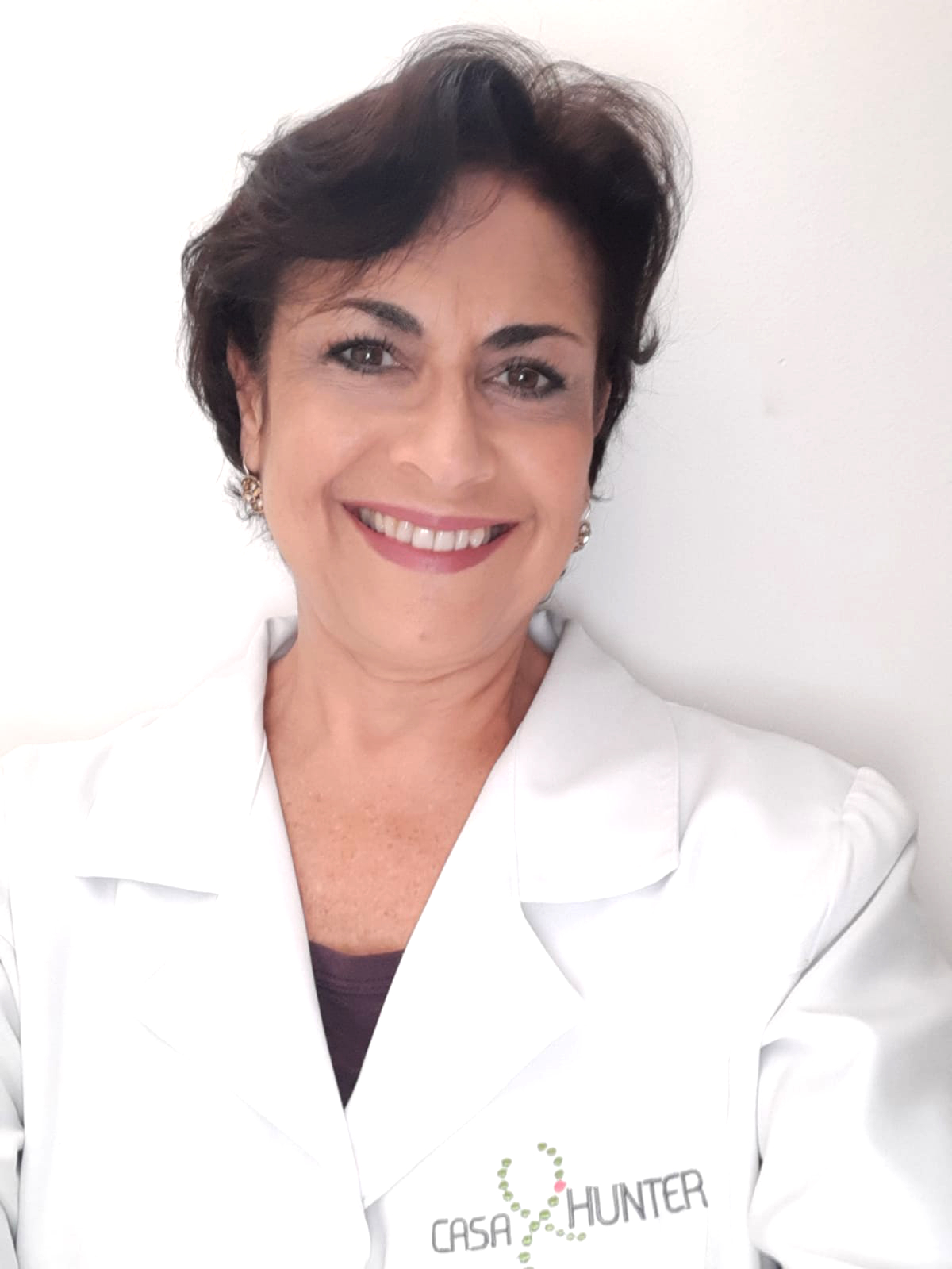 Foto da Regina Khoury. Ela posa para a foto e sorri. Regina tem cabelos castanhos curtos, usa blusa preta com jaleco branco com o logo da Casa Hunter. 