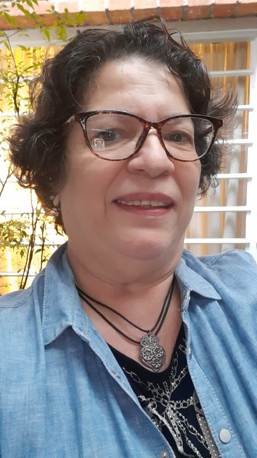 Foto da Idely Lelot. Ela posa para foto e sorri. Idely tem cabelos escuros curtos, usa óculos de grau, um colar, uma blusa preta com jaqueta azul.