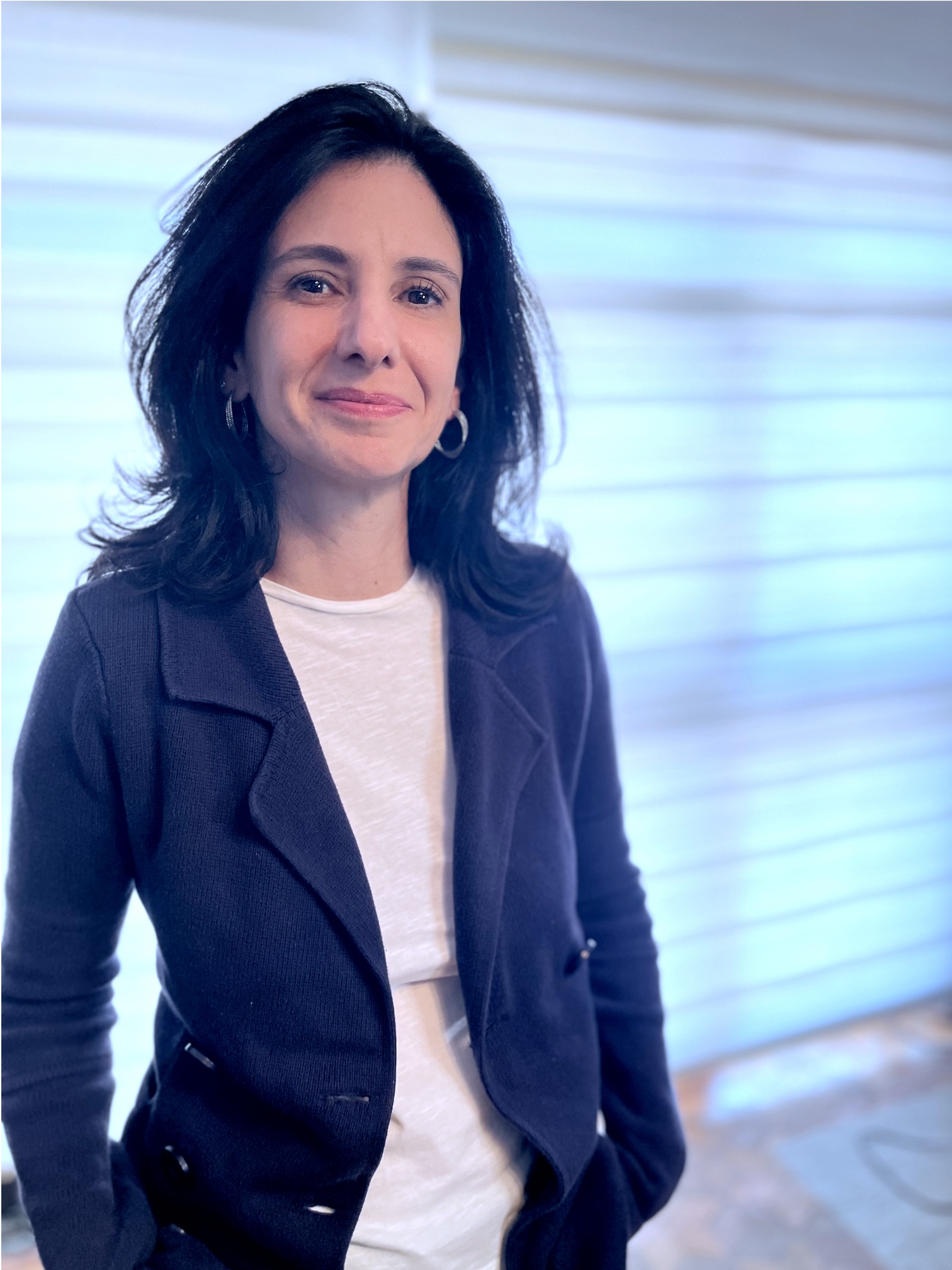 Foto da Juliana Barica Righini. Ela posa para a foto, está em pé e sorri. Juliana é uma mulher branca, tem cabelos castanhos escuros lisos, longos na altura dos ombros, usa uma blusa branca com um blazer azul marinho.