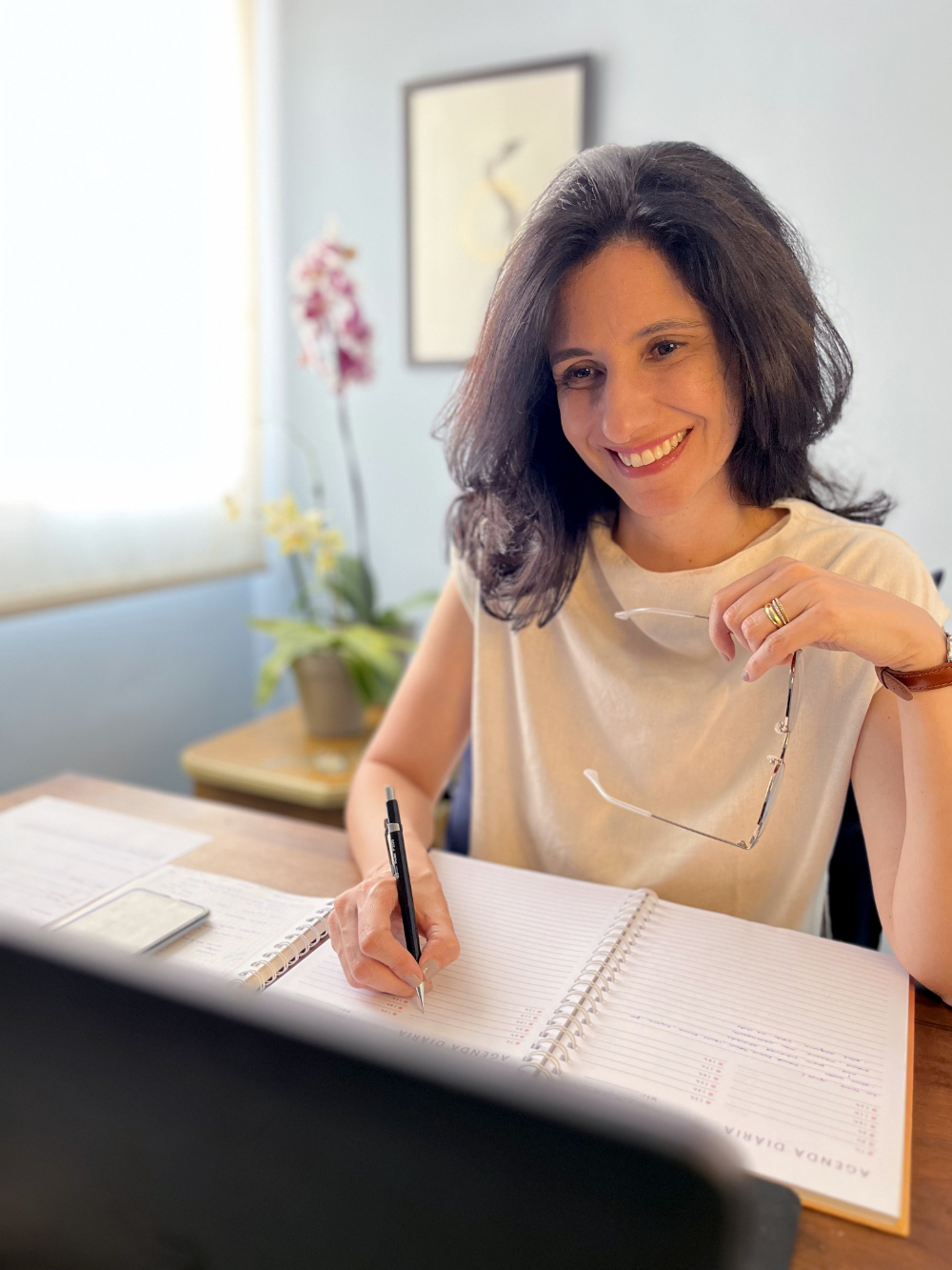 Foto da Juliana Barica Righini em um lugar fechado. Ela está sentada atrás de uma mesa. Juliana sorri e escreve em um caderno. Ela é uma mulher branca, tem cabelos castanhos, longos e lisos, que estão soltos. Usa uma blusa bege, sem mangas.