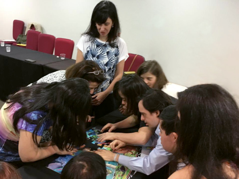 Foto da Juliana Barica Righini em pé ao lado de um grupo de pessoas em um lugar fechado. Elas estão reunidas ao redor de uma mesma atividade. Ao fundo, há algumas pessoas sentadas.