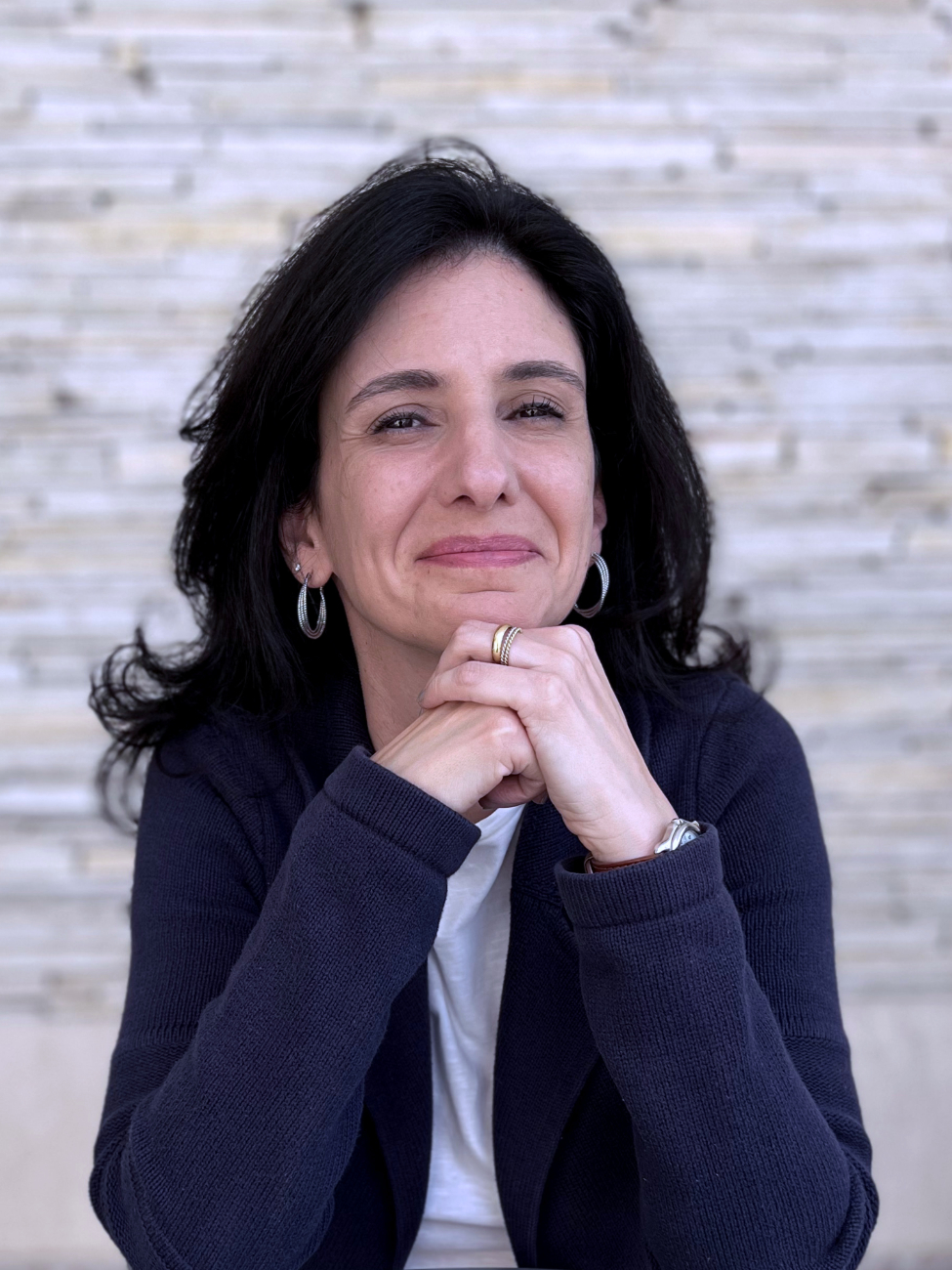 Foto da Juliana Barica Righini. Ela posa para a foto, sorri e está com as duas mãos unidas, apoiadas em uma mesa e próximas ao queixo. Juliana é uma mulher branca, tem cabelos castanhos escuros longos e lisos, usa brincos, uma blusa branca e uma jaqueta azul por cima.