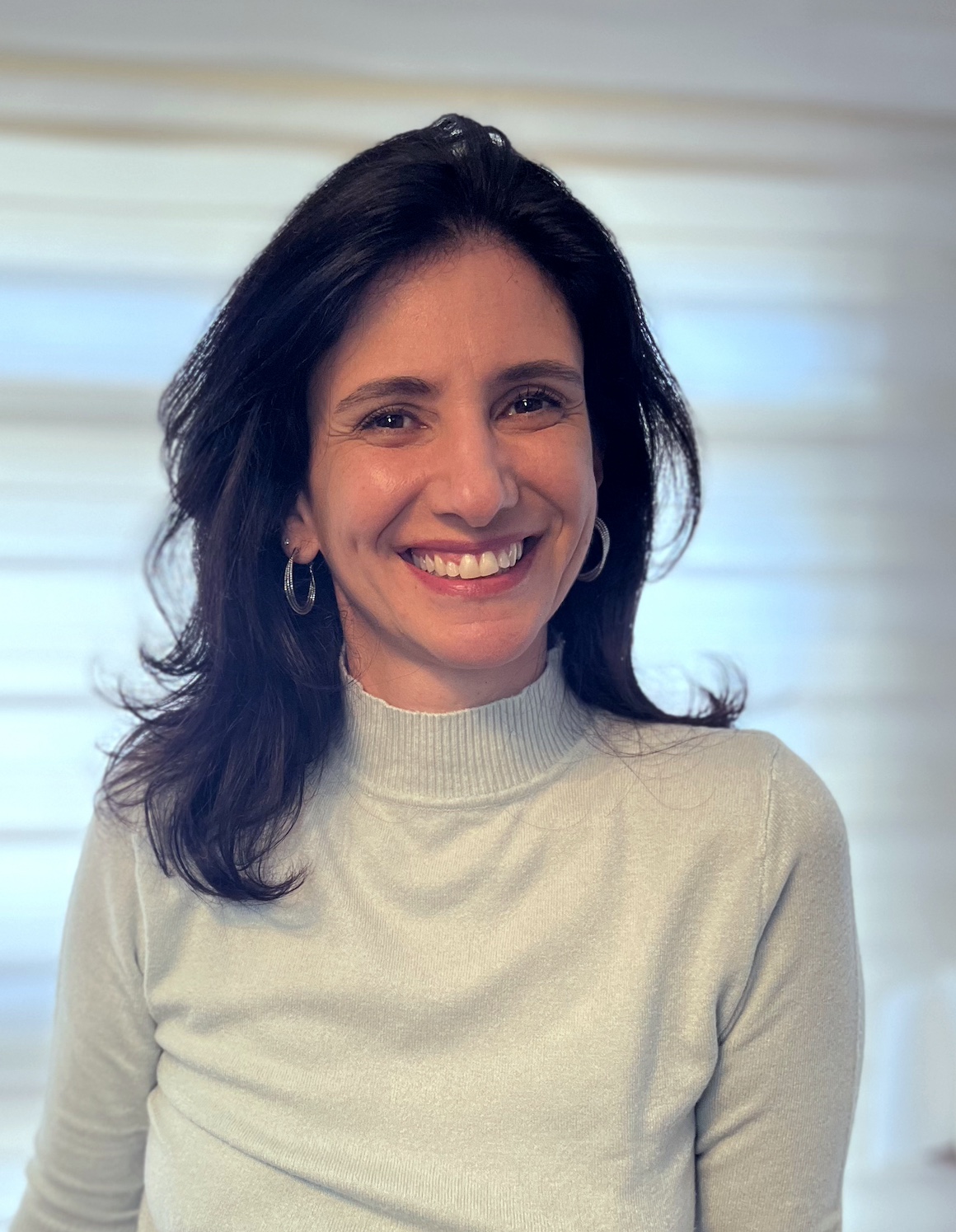 Foto da Juliana Barica Righini em pé em um lugar fechado. Ela posa para a foto e sorri. Juliana é uma mulher branca, tem cabelos castanhos escuros, lisos e longos abaixo dos ombros, usa brincos e blusa azul claro de gola alta com mangas compridas.