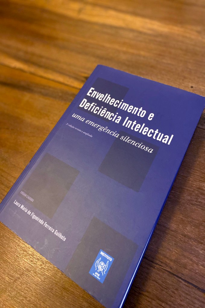 Foto do livro com capa azul marinho sobre uma mesa de madeira. O livro com título em branco "Envelhecimento e Deficiência Intelectual- uma emergência silenciosa." Na parte inferior o nomes da Laura Maria em branco e o logo da APAE de São Paulo.