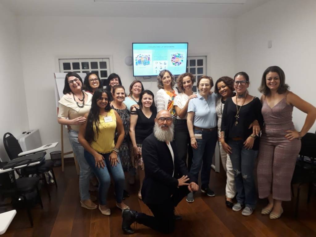 Foto de 14 pessoas reunidas em uma sala. Elas posam e sorriem para a foto. Há 13 mulheres em pé e um homem agachado ao centro. Há algumas cadeiras escolares ao redor. Juliana Barica Righini e Regiane Silva estão entre elas. 